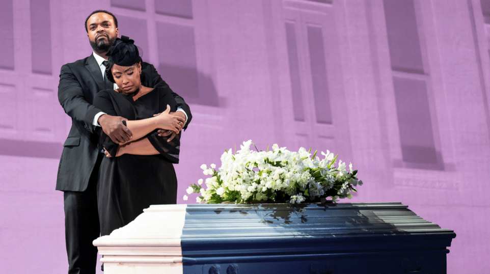 A tall African-American man hugs a shorter African-American woman from behind. The woman is crying. The man is looking off into the distance. They are standing by a white, closed casket with flowers on top.