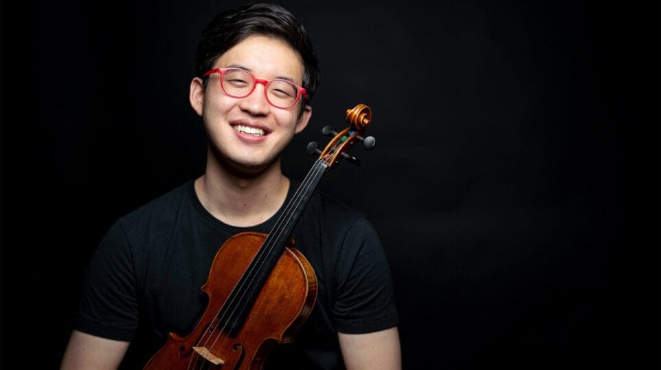 Julian Rhee smiles wearing red eyeglasses and black tee shirt swhile holding a violin