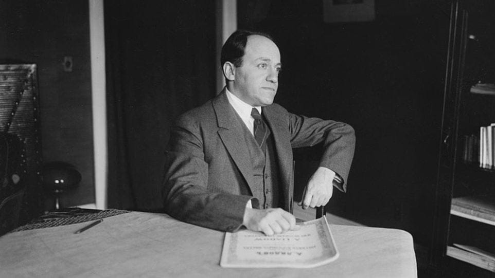 Ernest Bloch, seated at a table, looks into the distance while touching a score