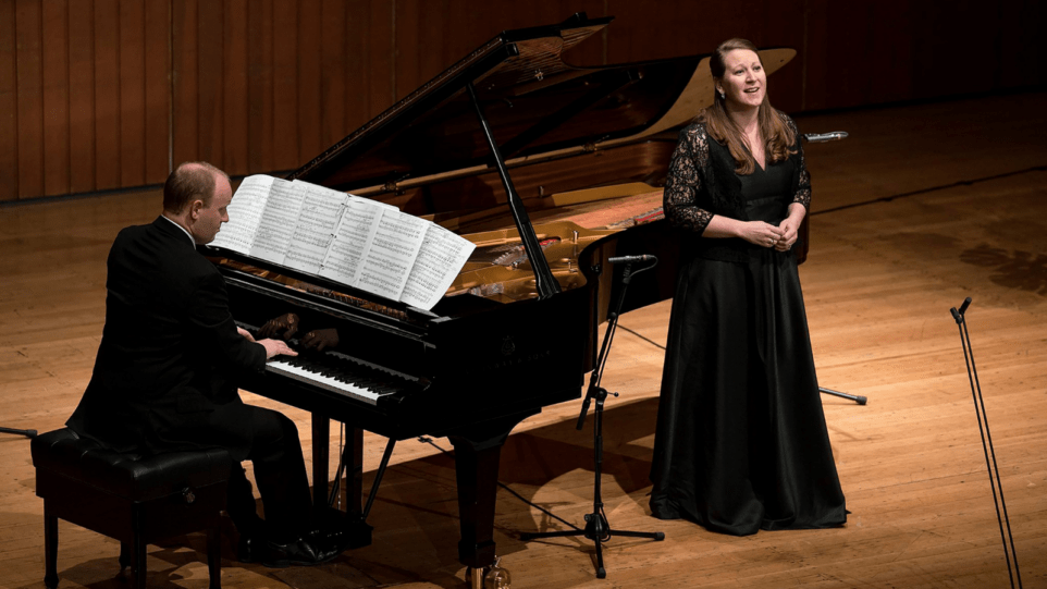 Clara Osowski and Tyler Wottrich onstage at the 2017 Das Lied Competition. Clara in black gown standing in crook of piano.
