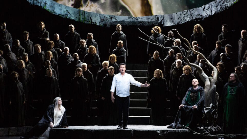 A scene from the Met's production of 'Lohengrin' — Piotr Beczała, in a white shirt and black trousers, sings standing among a chorus dressed in black, with Tamara Wilson, in white, seated to the left and Günther Groissböck and Brian Mullignan, both in black and green, to the right
