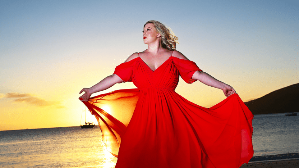 Laura Strickling in a flowing red gown on a beach at sunset