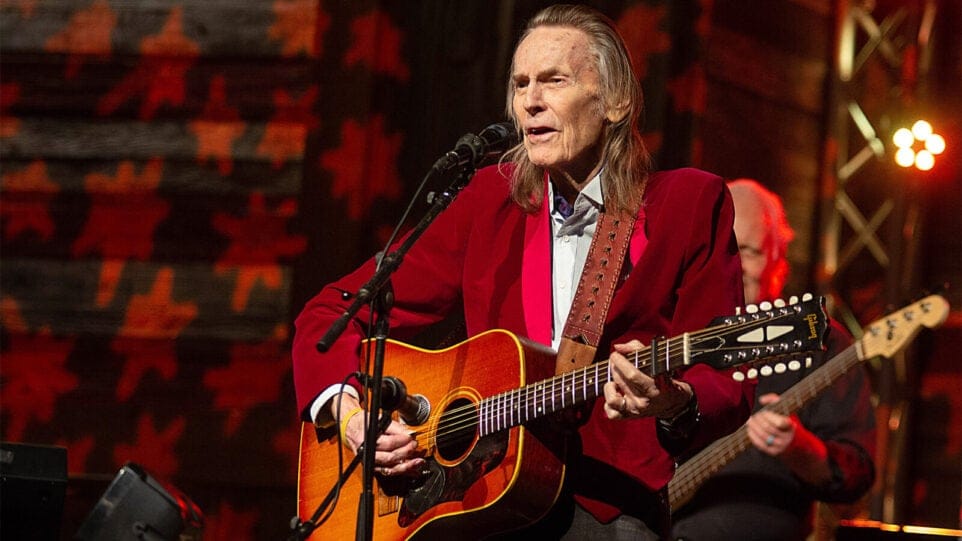 Gordon Lightfoot, in a deep red blazer, strums an acoustic guitar and sings into a mic