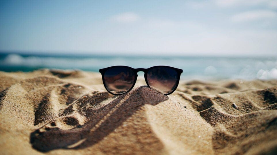 sunglasses sit on a sandy beach with water in the background
