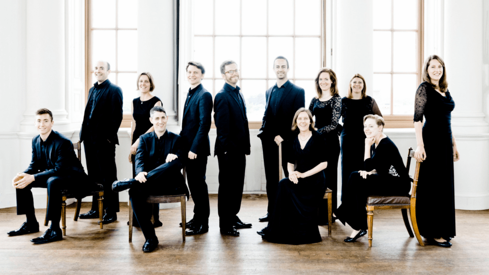 The twelve members of the ensemble in concert black attire posing in front of a large picture window through which a lot of light is coming; white walls, hardwood floors.