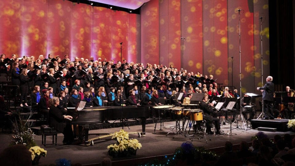 a stage filled with chorus members performs against a festive red backdrop