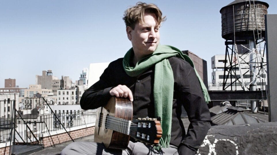 Jason Vieaux, sporting a black shirt, green scarf, and holding a guitar, sits on an urban rooftop and smiles