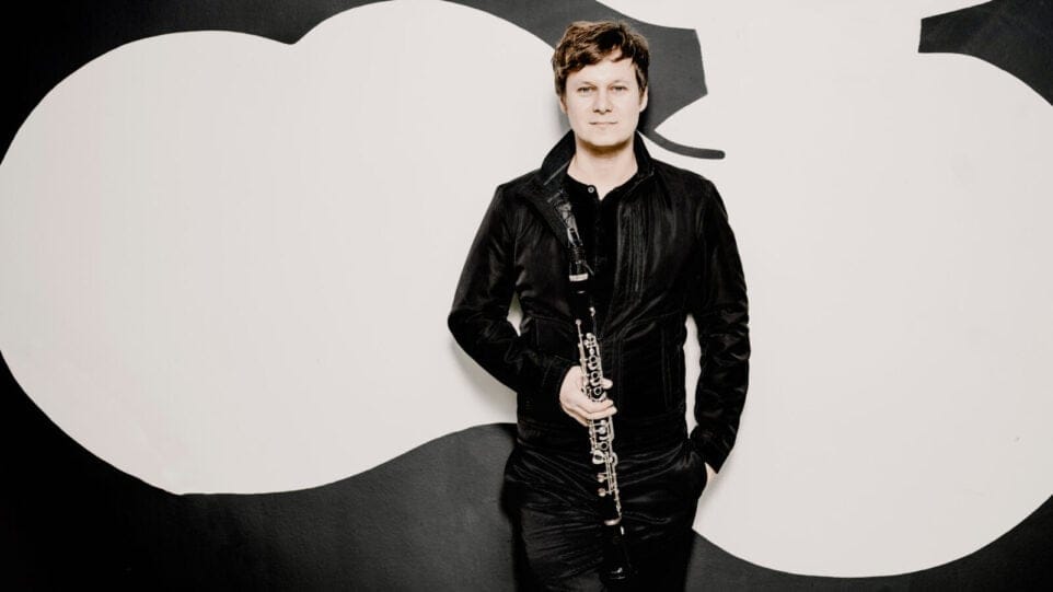 Sebastian Manz holds a clarinet, leaning against a striking black and white geometric background
