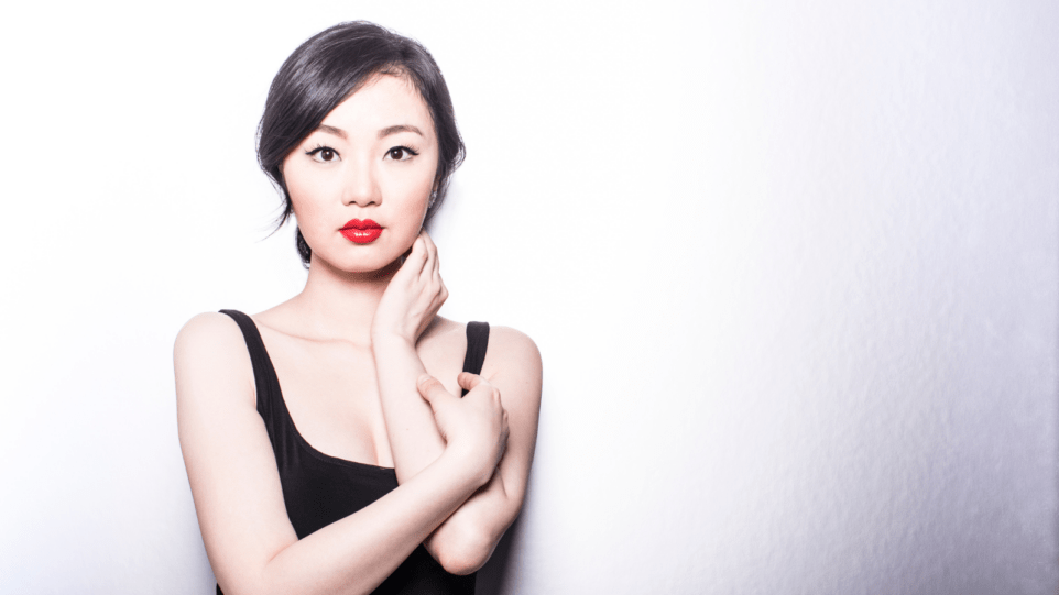 Ying Fang wearing a sleeveless black dress, red lipstick, left hand touching her neck, right hand holding left forearm, white background creating strong contrast.