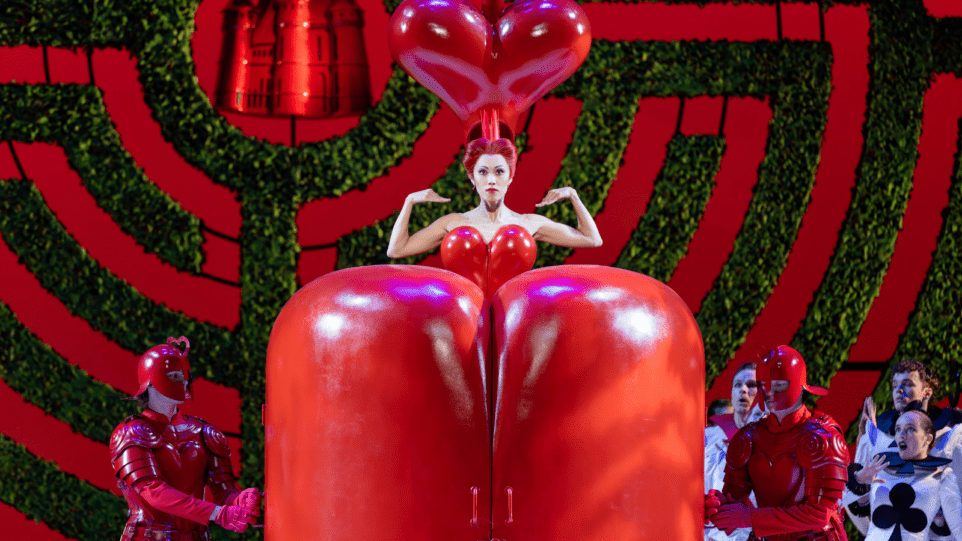An abnormally tall woman dressed in a red, heart-shaped dress, has a red, heart-shaped headdress that appears to be floating over her head. It is unclear if she is sitting on top of two rounded red columns that look like a door, or if those two rounded red columns are supposed to be the character's legs.
