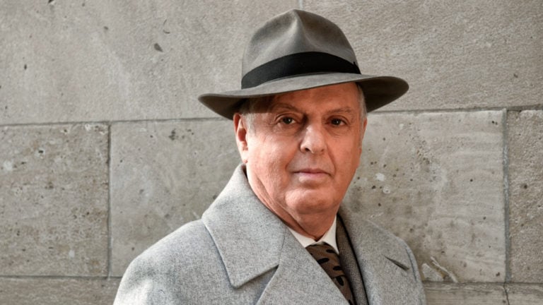 Daniel Barenboim in a grey hat, stands in front of a stone wall