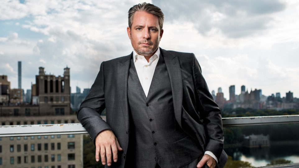 Matthew Polenzani in a grey suit and white shirt poses on a balcony against a sunny skyline