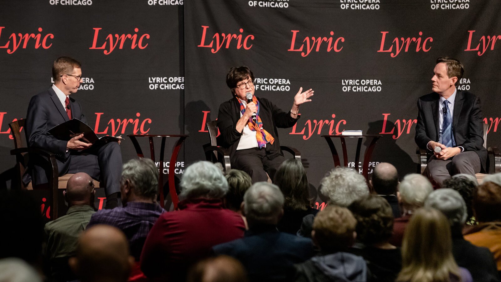 onstage at a Lyric Opera panel, Sister Helen Prejean speaks, center, with George Preston and Jake Heggie looking on thoughtfully. A full audience observes attently