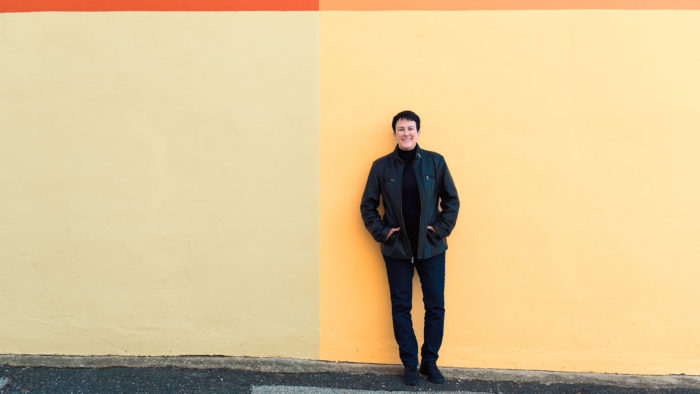 conductor Jennifer Higdon in jeans, a black turtle neck, and black leather jacket standing in front of a simple mural of four colors: red, orange, yellow, and beige.
