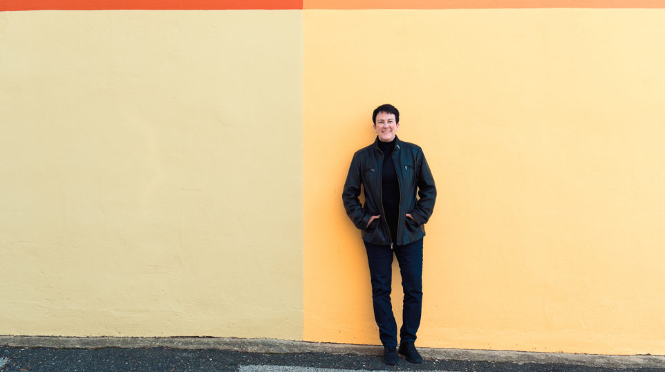 conductor Jennifer Higdon in jeans, a black turtle neck, and black leather jacket standing in front of a simple mural of four colors: red, orange, yellow, and beige.