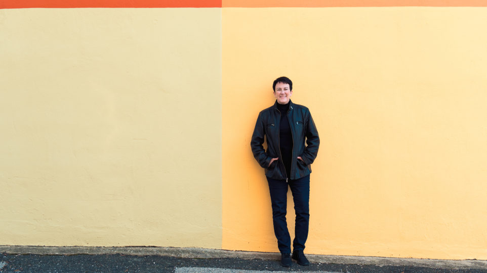 conductor Jennifer Higdon in jeans, a black turtle neck, and black leather jacket standing in front of a simple mural of four colors: red, orange, yellow, and beige.
