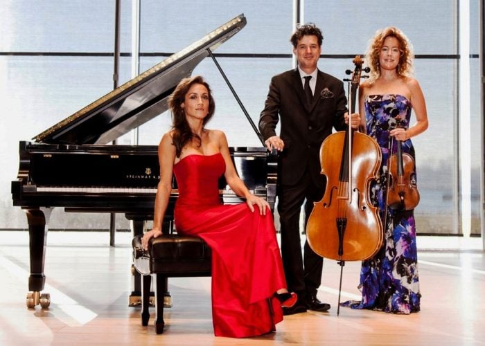 three formally dressed musicians pose in front of windows with a view of water and a skyline