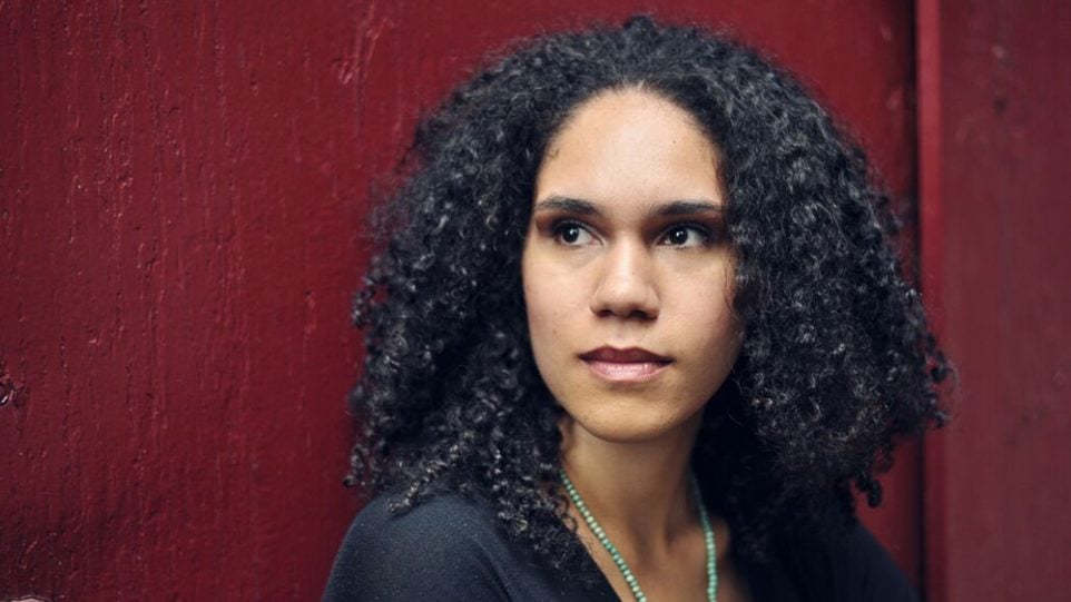 wearing a green necklace and a black sweater, Jessie Montgomery looks past the camera standing against a rich red wall