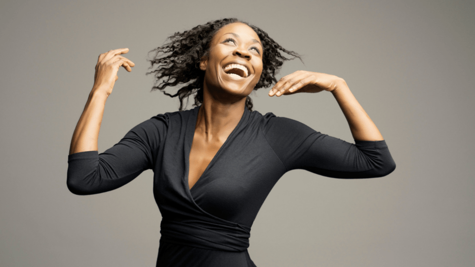 Portrait of Jeanine De Bique in tailored balck dress with long sleeves, whipping her hair to the side.