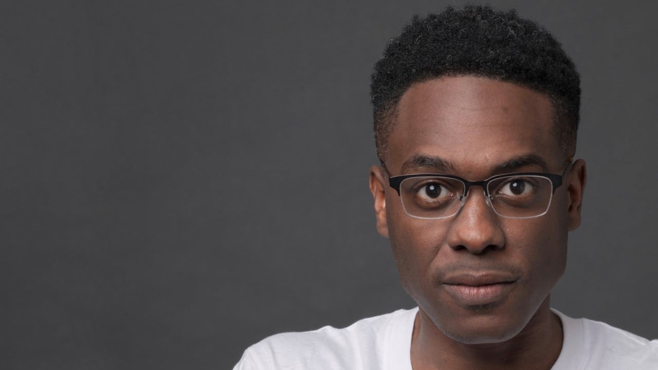 Headshot of Shawn Okpebholo, looking straight at camera wearing black half-frame glasses and white t-shirt.,