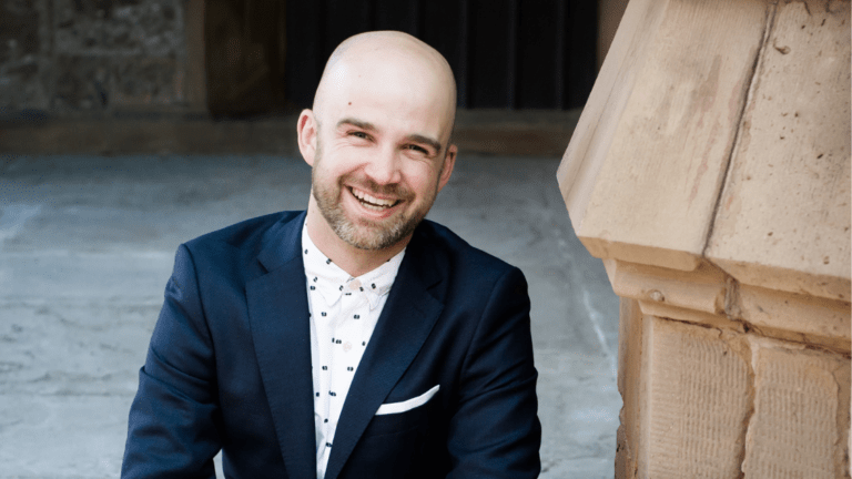 Portrait of Zachary WIlder seated on stone staircase, blue suit, white shirt no tie, smiling widely