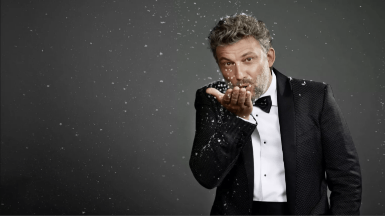 Jonas Kaufmann in a tuxedo blwoing snow off his hand toward the camera