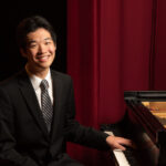 Tyler Wang, a young pianist, poses at the keyboard with a red curtain in the background