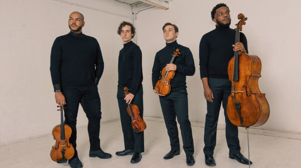 Four young men wear black, pose with their instruments in a white studio