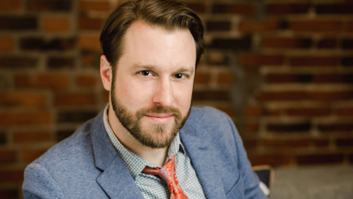 Portrait of Jesse Blumberg in light blue suit, red brick wall out of focus in background