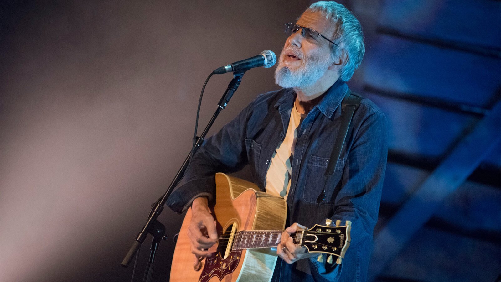 Cat Stevents, wearing sunglasses, a denim shirt, and a cream colored tee shirt, strums a guitar and sings into a microphone onstage