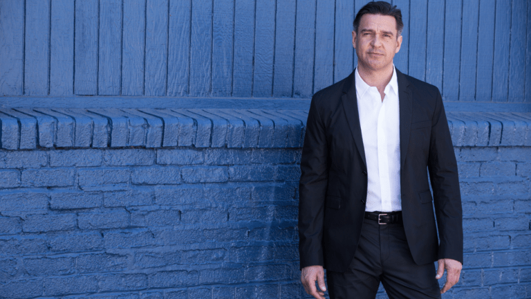 Color portrait of nathan Gunn wearing dark suit, white shirt, no tie, in front of brick wall painted blue.