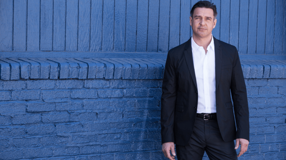 Color portrait of nathan Gunn wearing dark suit, white shirt, no tie, in front of brick wall painted blue.