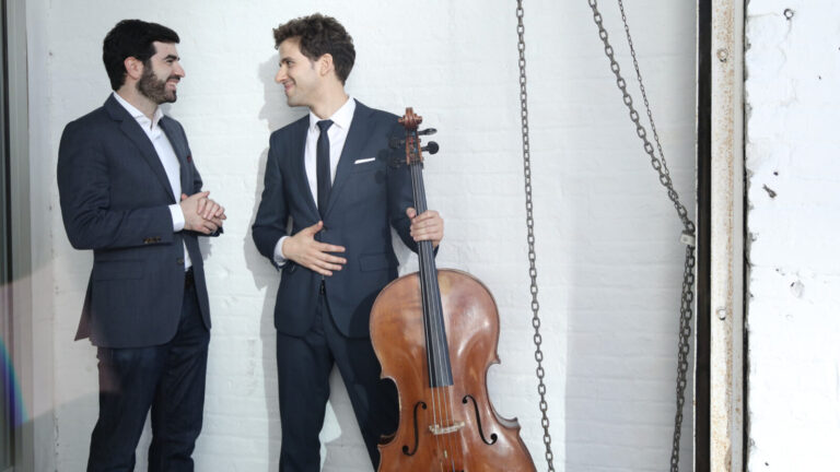 Michael Stephen Brown and Nicholas Canellakis, dressed in suits, share a laugh in front of a white brick wall