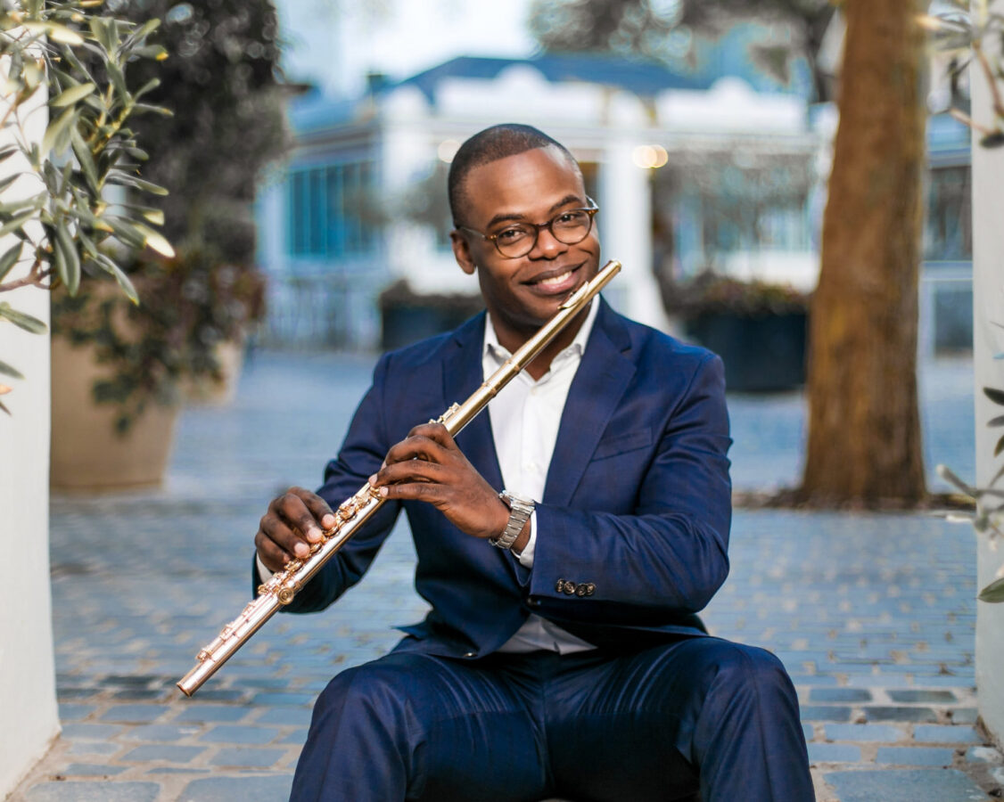 Demarre McGill, seated outside, smiles at the camera while holding the flute