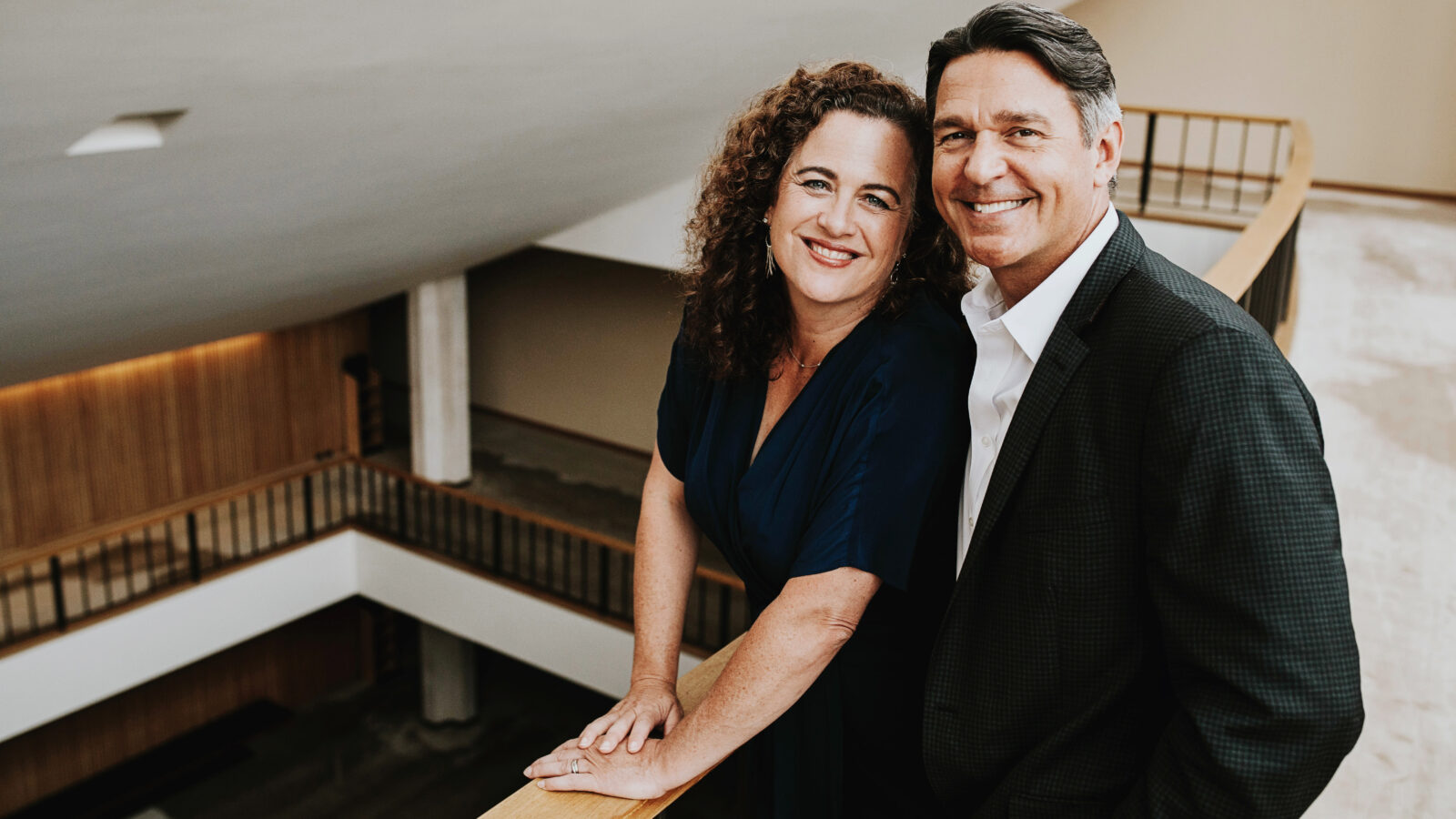 Color portrait of Julie Jordan Gunn in a dark blue dsress and Nathan Gunn, in a dark suit and no tie, posing in a modern lobby