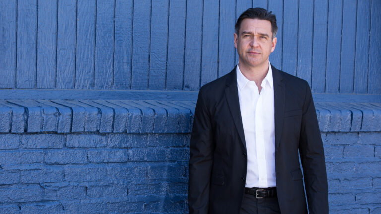 Color portrait of nathan Gunn wearing dark suit, white shirt, no tie, in front of brick wall painted blue.