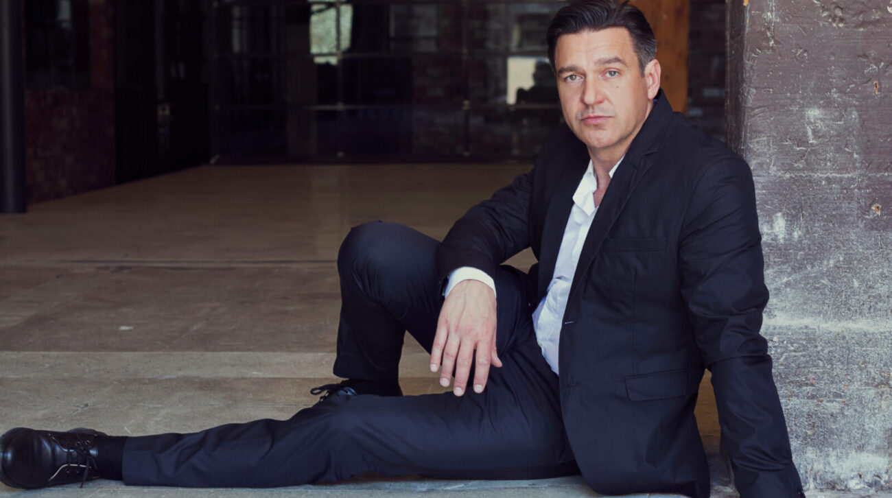 Color portrait of nathan Gunn wearing dark suit, white shirt, no tie, seated leaning against a brick wall