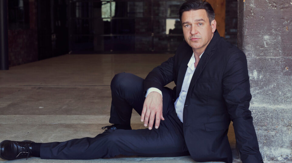 Color portrait of nathan Gunn wearing dark suit, white shirt, no tie, seated leaning against a brick wall