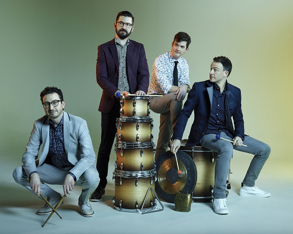 Third Coast Percussion poses with percussion instruments and mallets in front of a yellow-green background