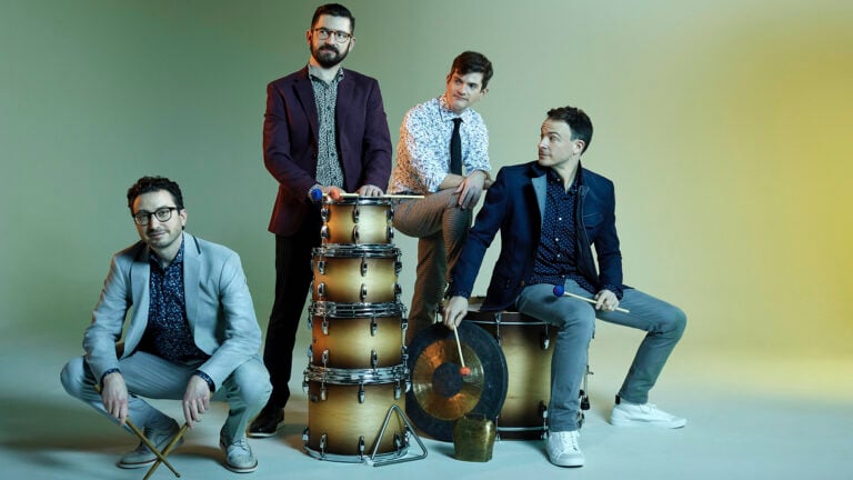 Third Coast Percussion poses with percussion instruments and mallets in front of a yellow-green background