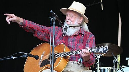 white haired man with hat points to his right while holding guitar