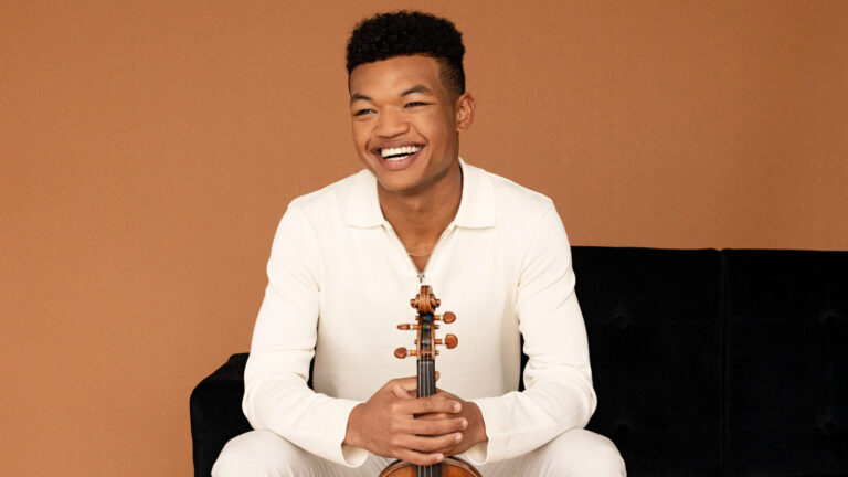 Violinist Randall Goosby sits with his instrument on a black couch against a tan background