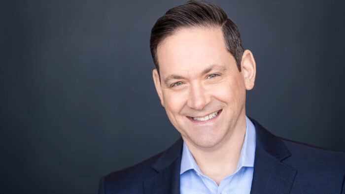 Lawrence Edelson, wearing a blue checked suit and a blue dress shirt, smiles against a grey-blue background