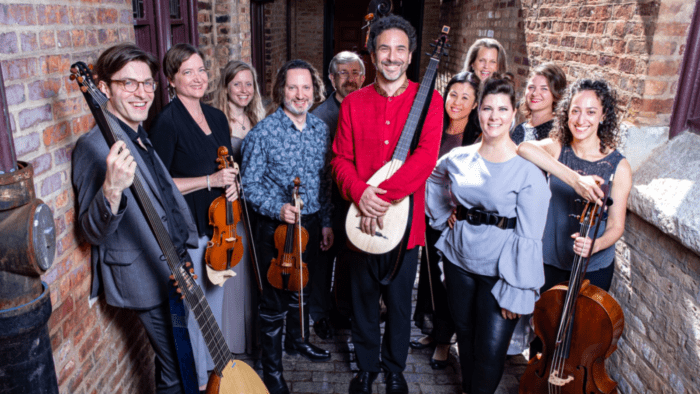 Third Coast Baroque: 11 musicians in a sunlit red brick hallway; some with their instruments including violins, cello, theorbo, and charango.