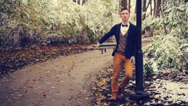 July 7 - July 11, 2025 Man in cardigan and bowtie holds model piano and leans against a park lightpost
