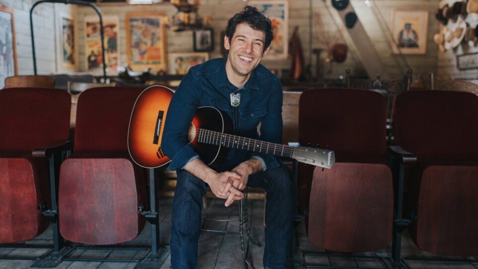 Man in western shirt with guitar sits in rustic theatre