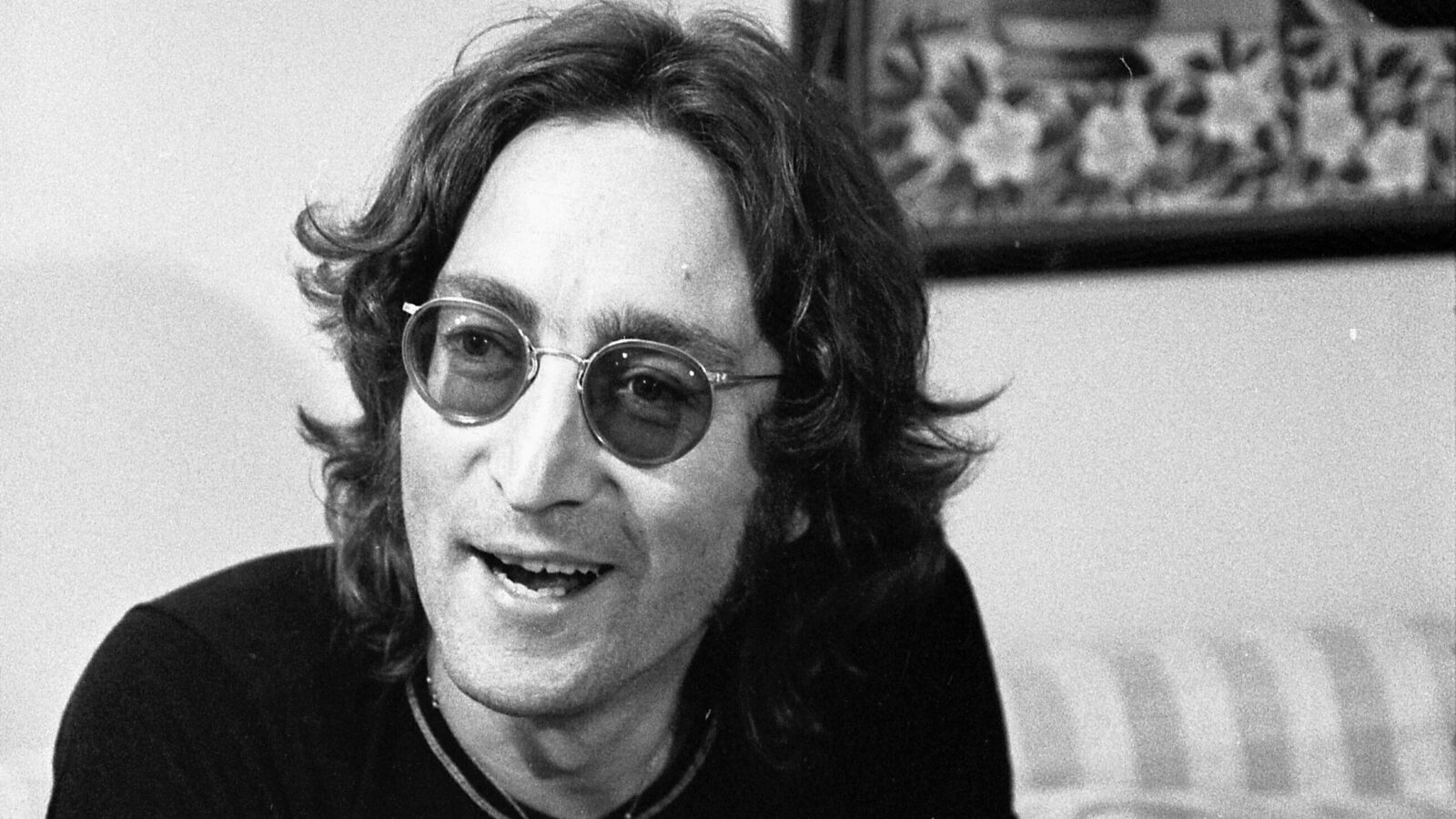 John Lennon, with long hair and his signature round glasses, smiles