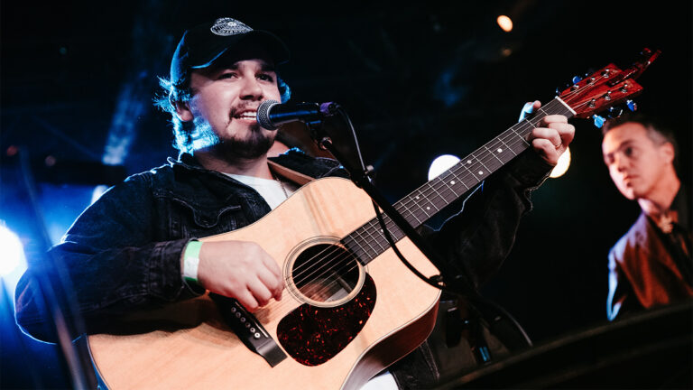 a man in a jean jacket, trucket hat, and white shirt — Tommy Prine — strums a guitar and sings into a microphone onstage