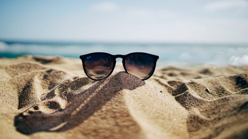 sunglasses sit on a sandy beach with water in the background