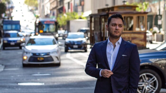 Nicholas Phan in grey suit, white shirt, no tie standing in an urban road, traffic moving toward the camera